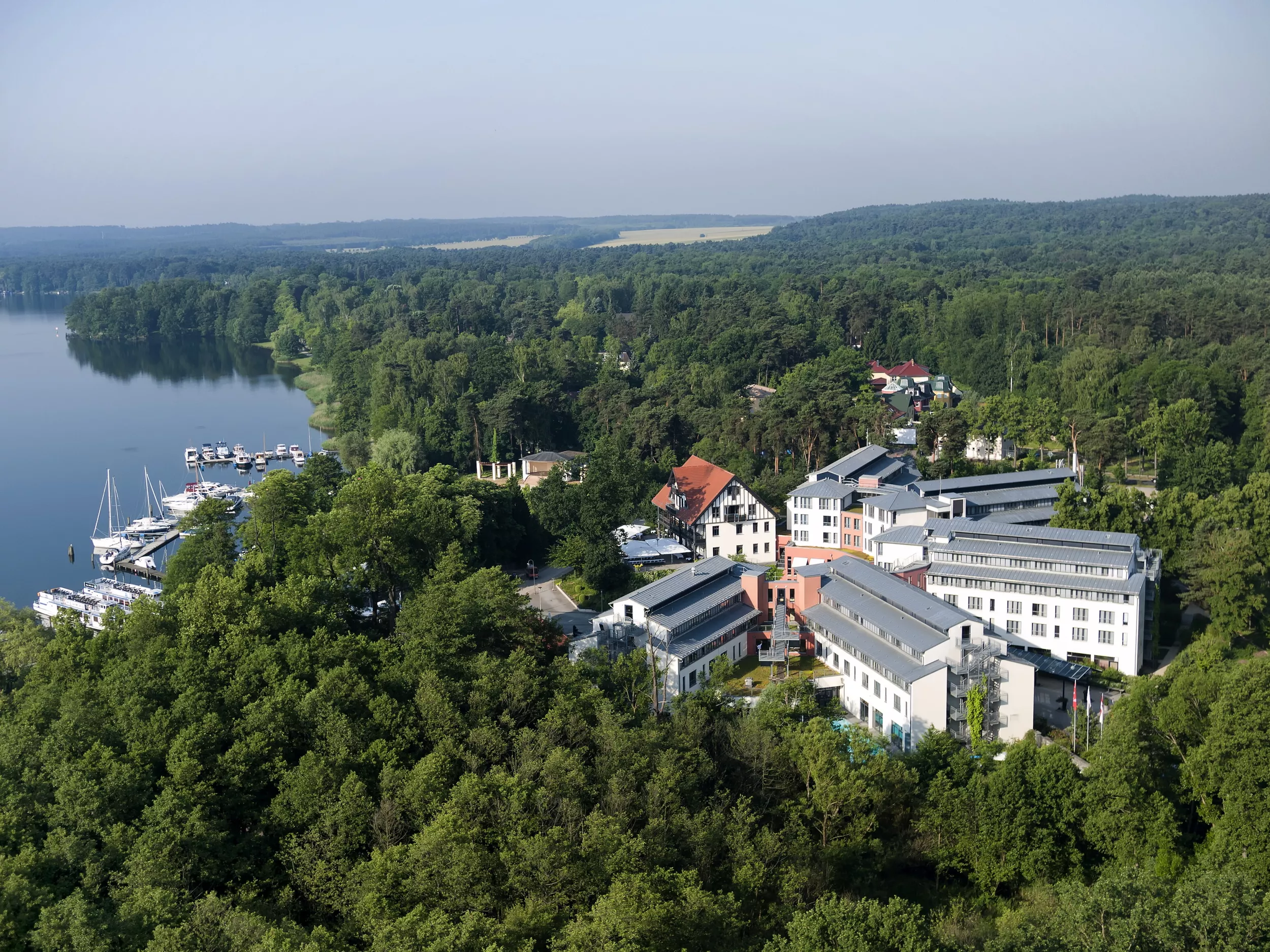 Aufnahme von der Luft auf Hotel, links beginnt der See mit Anliegestelle für Schiffe und Boote. Hotel von grünen Bäumen umgeben