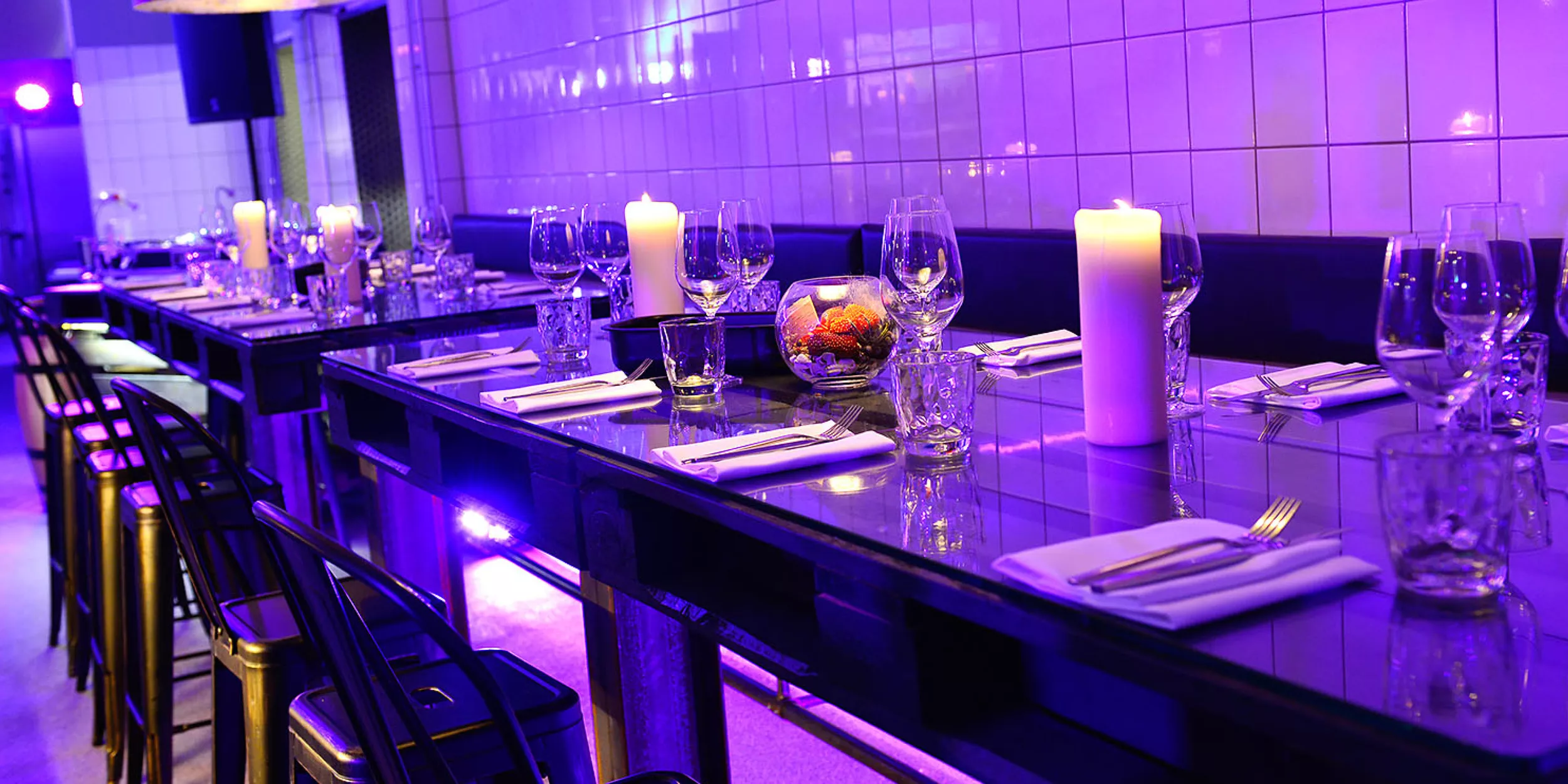Gedeckter Paletten-Tisch mit Glasplatte, Gedecke mit Besteck, Gläsern und Blumendekoration in einer stylisch lila beleuchteten Hotelküche.