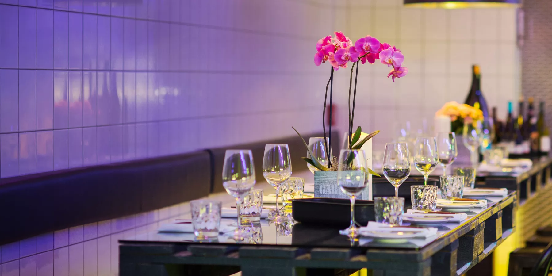 Gedeckter Paletten-Tisch mit Glasplatte, Gedecke mit Besteck, Gläsern und Blumendekoration in einer mit gelb und lila beleuchteten Hotelküche.