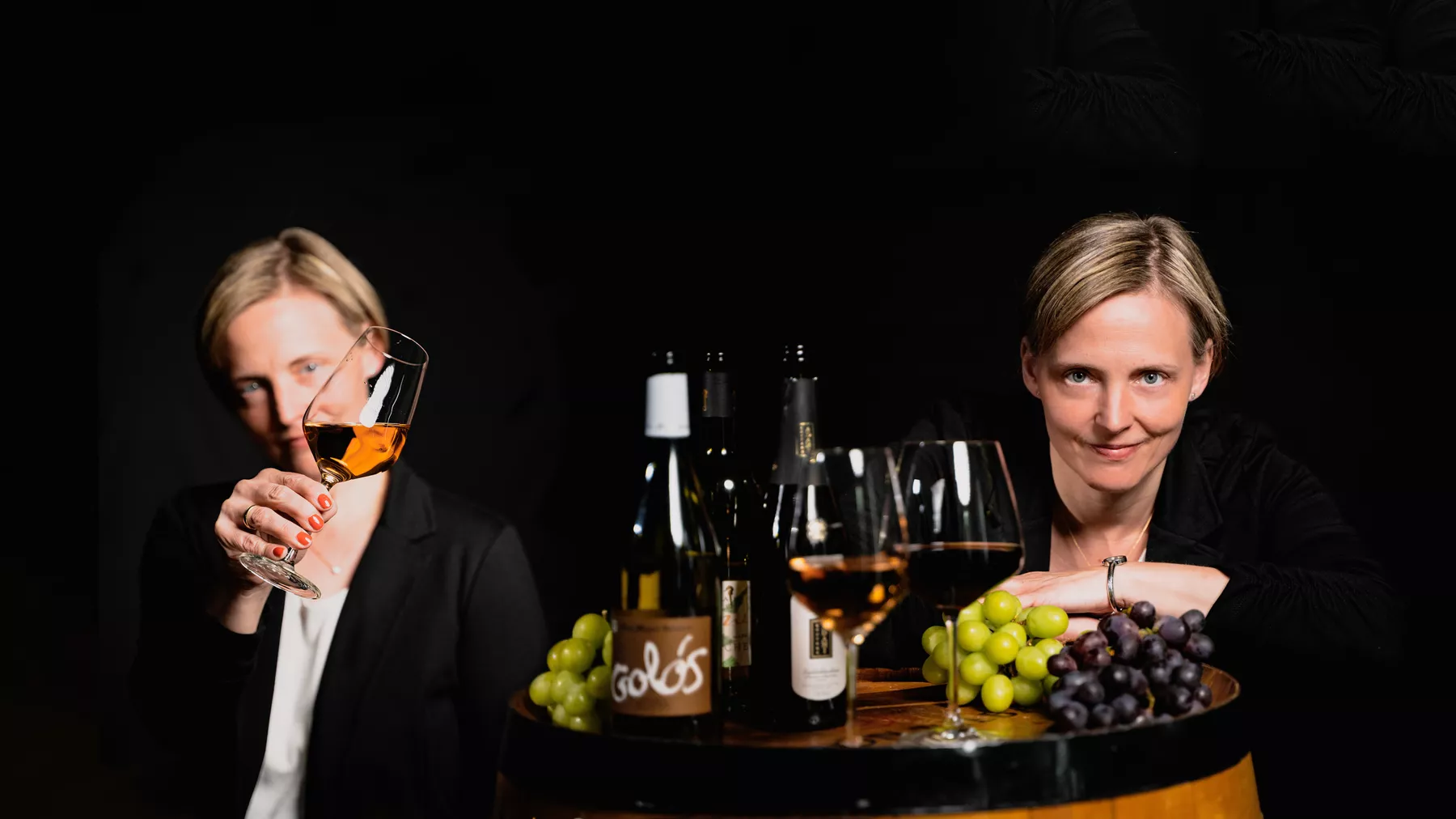 gleiche Frau steht links mit schräg gehaltenem Weinglas und rechts hinter dem Weinfass und lächelt in die Kamera, auf dem Fass stehen zwei Weingläser, Weinflaschen und Weintrauben
