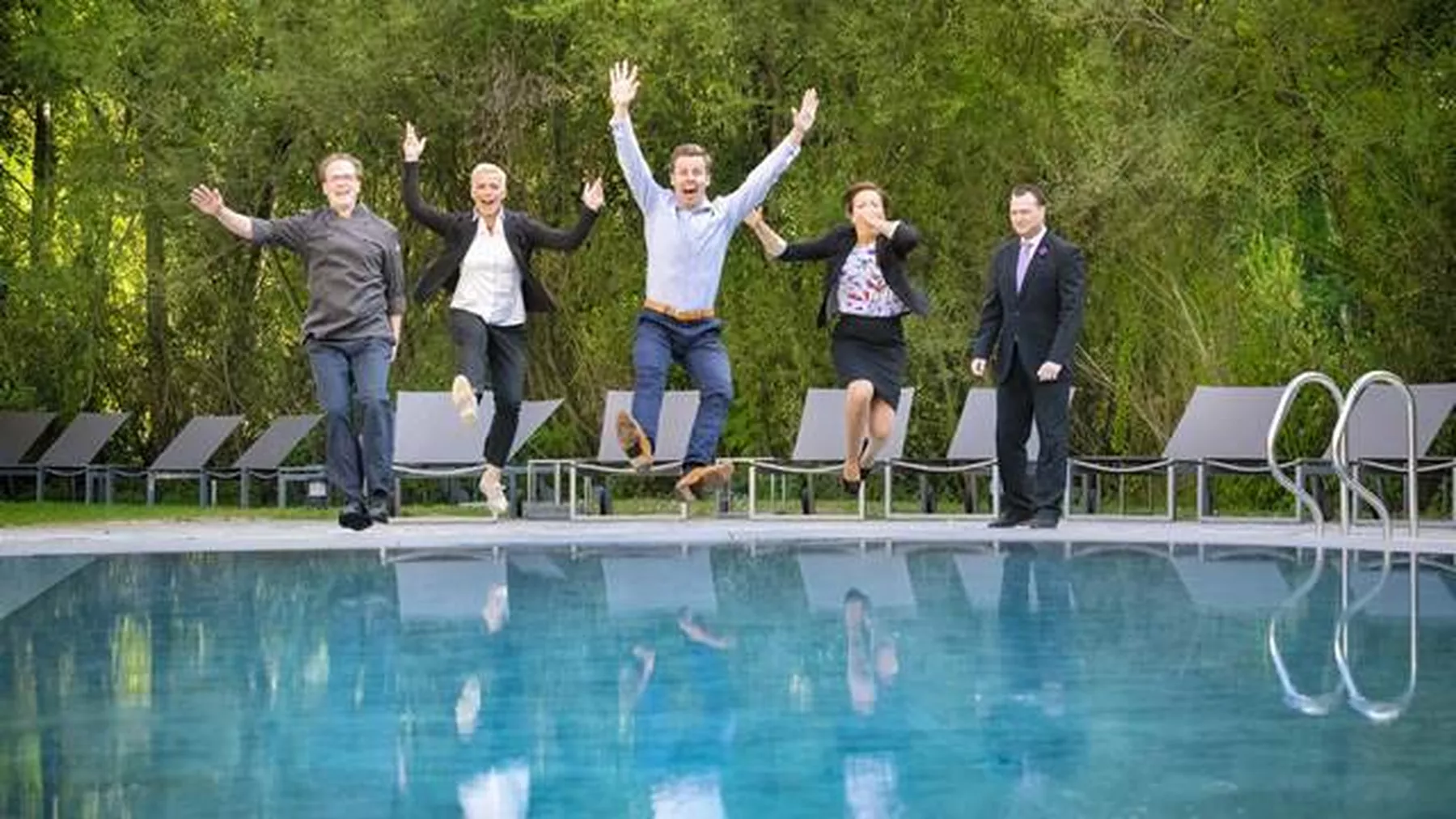 fünf Männer und Frauen springen in Arbeitskleidung freudig in den Pool, dahinter Liegen und grüne Stäucher